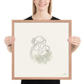 First Kiss, a fine-line drawing of a mother kissing her baby, Beloved print by Jamie Lawrence in a Red Oak frame, held to show scale